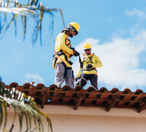 Two construction workers in safety gear working on a tiled roof under a clear blue sky.